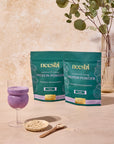 Two packages of Neeshi protein powder beside a glass of protein shake and a small bowl of oats, with a plant in the background.